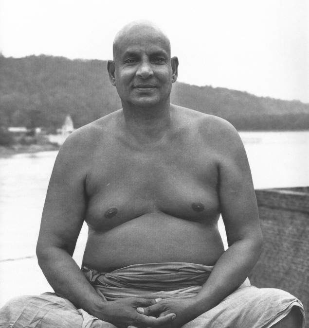 Swami Sivananda sitting by the Ganges smiling with a strong gaze