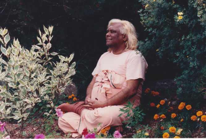 Swami Vishnu sitting cross legs smiling in nature next to flowers and bushes