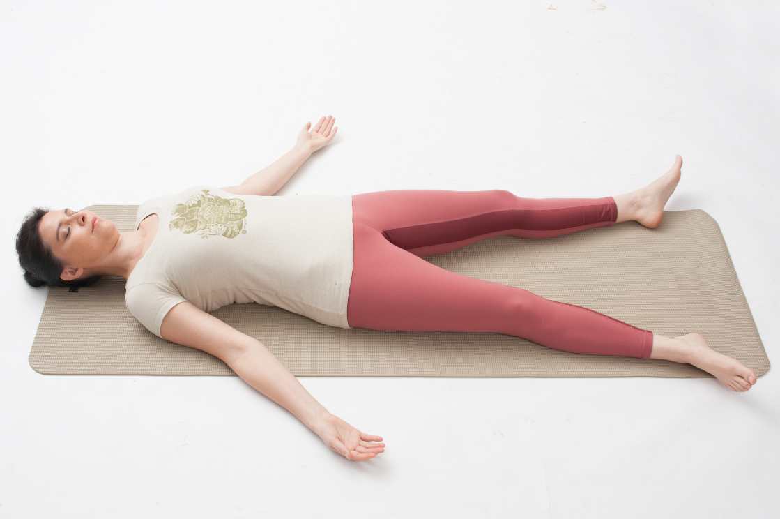 Student laying in Savasana - corpse pose, on her back, arms resting gently at 45 degree angle, palms up, and legs resting comfortably apart