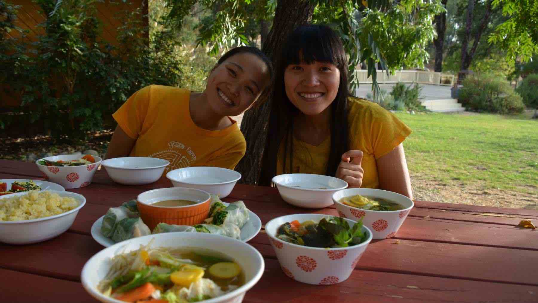 2 Female yogis smiling and enjoying lunch at the ashram.  Bowls of rice, soup, vegetables, spingrolls fill the picnic table.