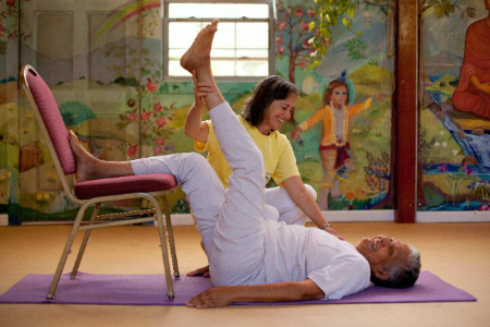 Woman is laying on a yoga mat on her back with one leg resting on the seat of a chair, and a teacher is helping to hold her other leg in the air for a gentle stretch.
