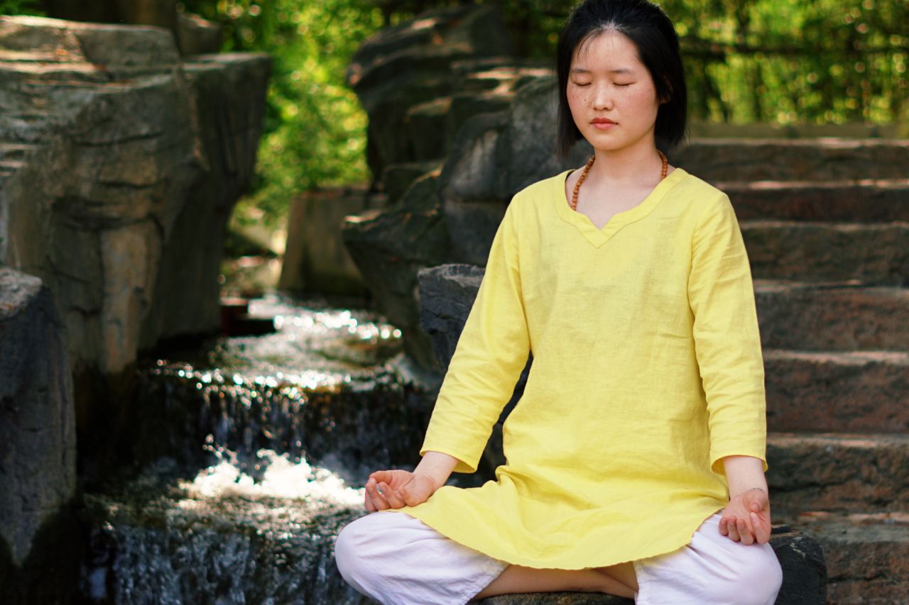 A Yoga Teacher Training student meditates next to outdoor stairs and a babbling brook.